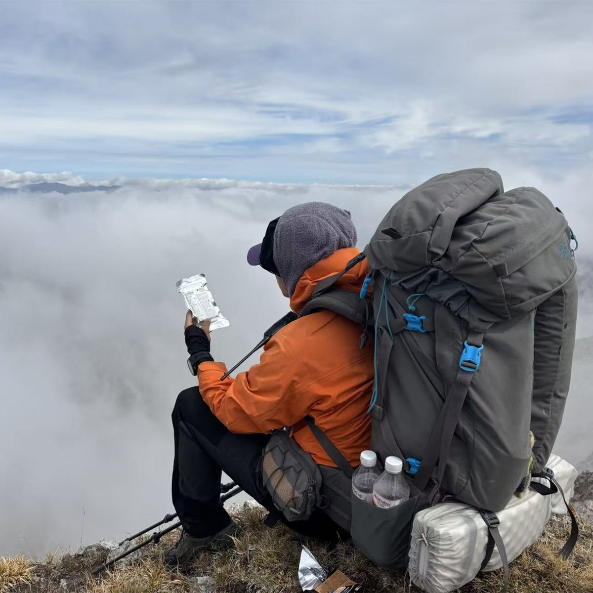 A hiker sitting on a mountain ridge above a sea of clouds, wearing an orange jacket, black pants, and a large gray backpack while holding PEAK4RATION EF Emergency food ration bar.