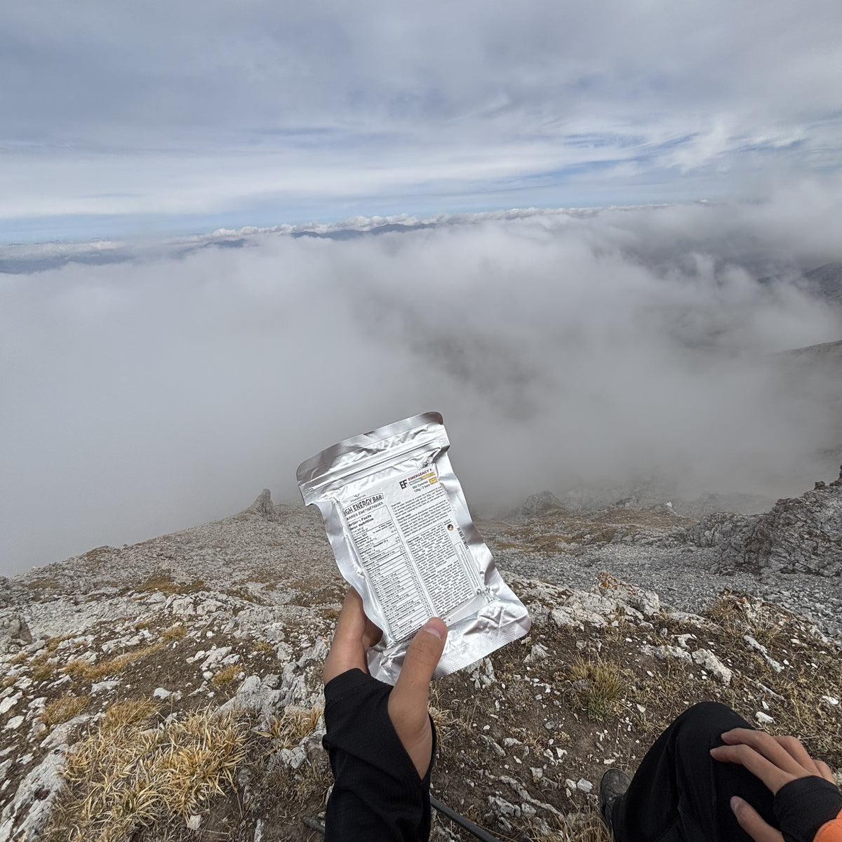 A person on a rocky mountain peak holds up PEAK4RATION EF Emergency food ration bar, showing the back of the package with printed text, with a sea of clouds and mountain landscape in the background.
