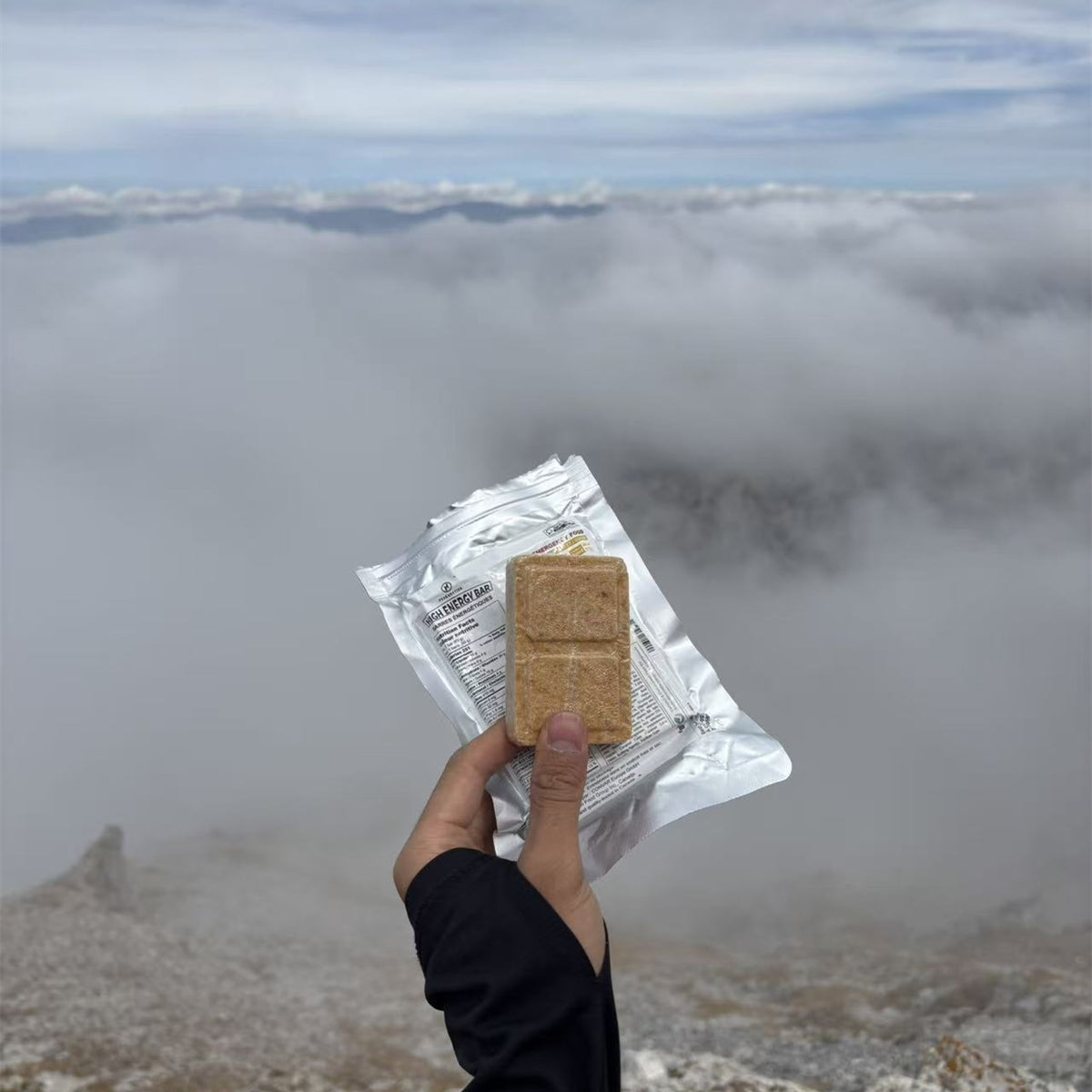 A hand holds a packaged snack bar in front of a misty mountain landscape with clouds and distant peaks. A hand wearing a purple glove holds a silver pouch of PEAK4RATION Energy Bar emergency ration food supply and a square piece of the golden-brown baked bar in front of a blurred outdoor mountain landscape.