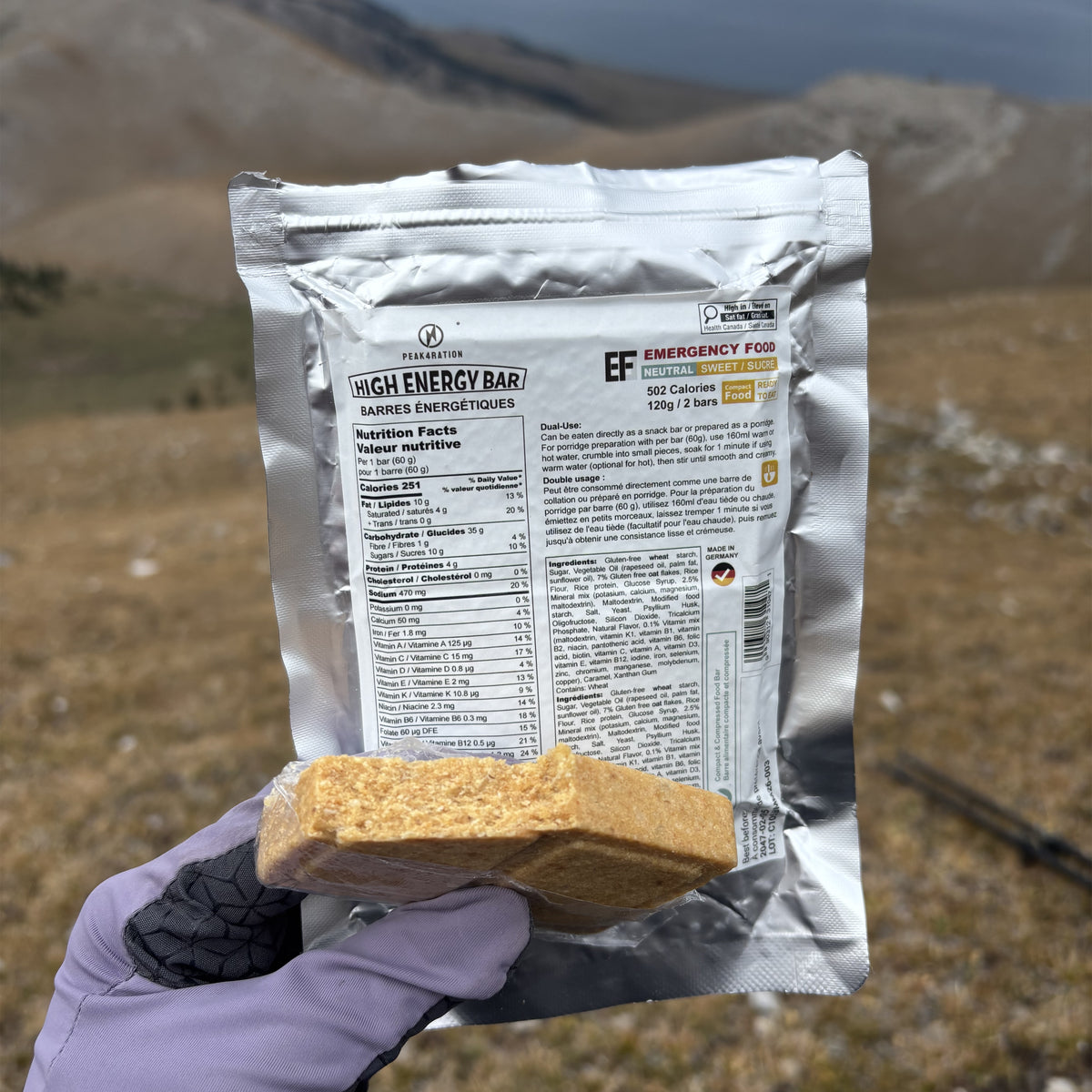 A hand wearing a purple glove holds a silver pouch of PEAK4RATION Energy Bar emergency ration food supply and a square piece of the golden-brown baked bar in front of a blurred outdoor mountain landscape.